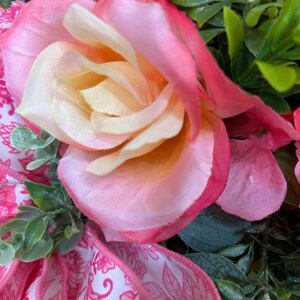 May include: A decorative arrangement featuring a large, multi-layered rose with petals transitioning from a pale cream center to vibrant pink edges. The rose is surrounded by green foliage and a white ribbon with a pink floral pattern.