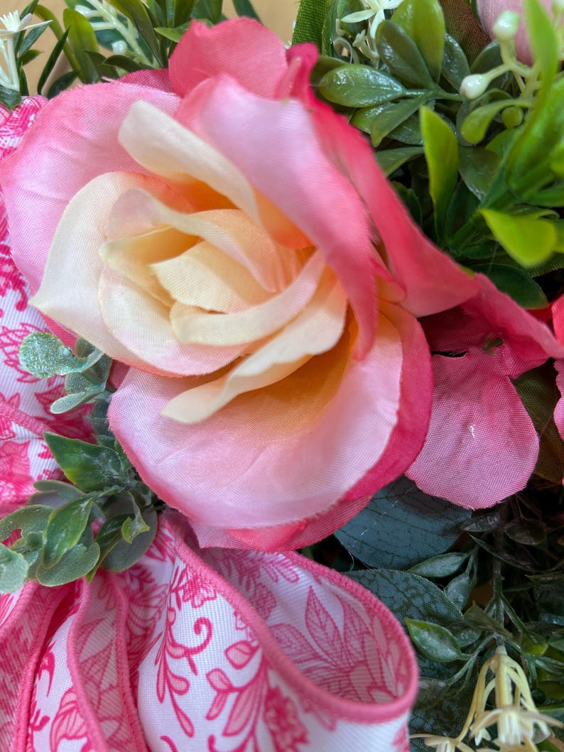 May include: A decorative arrangement featuring a large, multi-layered rose with petals transitioning from a pale cream center to vibrant pink edges. The rose is surrounded by green foliage and a white ribbon with a pink floral pattern.