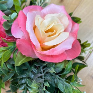 May include: A decorative wreath featuring a large central rose with cream-colored inner petals and vibrant pink outer petals. The arrangement includes various green foliage, small white filler flowers, and additional pink rose buds nestled within a woven base.