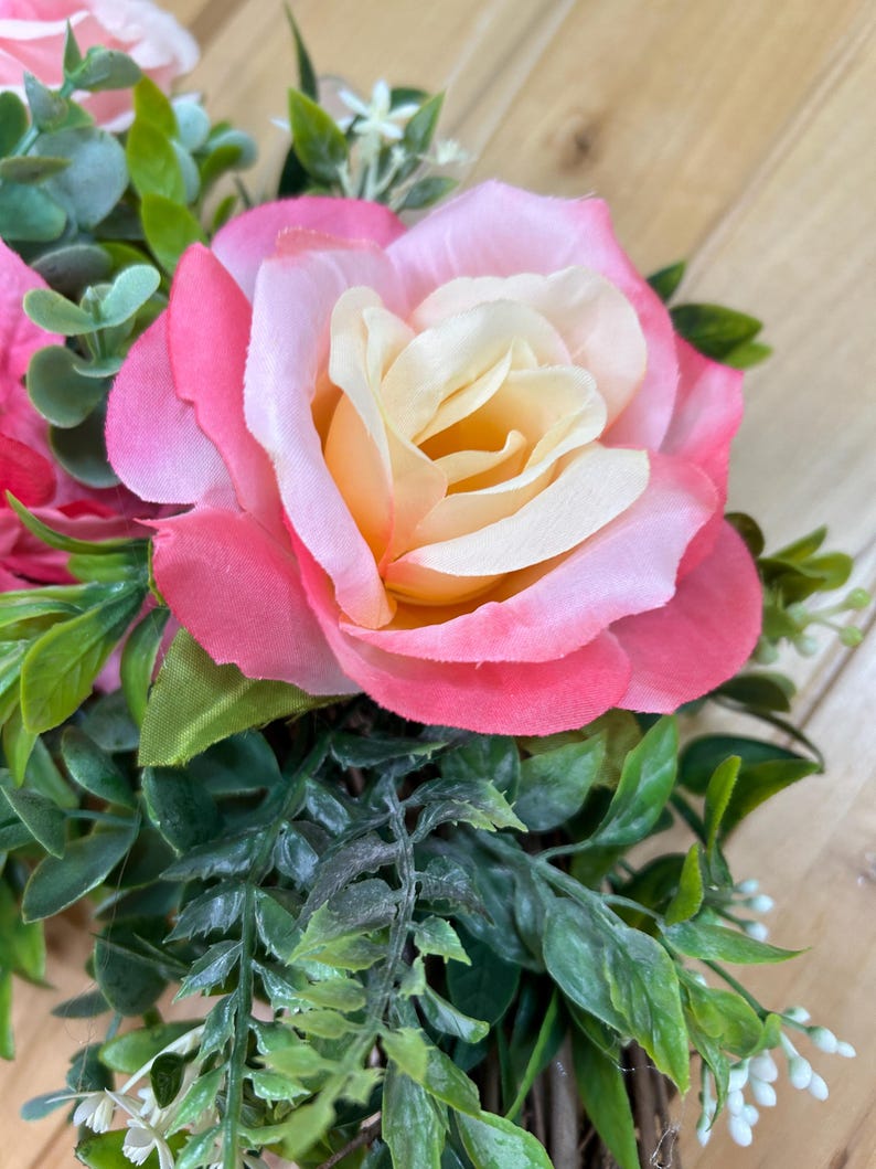 May include: A decorative wreath featuring a large central rose with cream-colored inner petals and vibrant pink outer petals. The arrangement includes various green foliage, small white filler flowers, and additional pink rose buds nestled within a woven base.