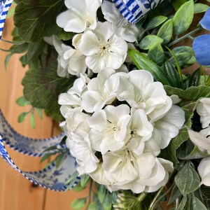 May include: A decorative wreath featuring clusters of white geranium flowers, green foliage, and a blue and white striped ribbon with a floral pattern. The arrangement includes various leaves and stems interspersed among the white blossoms.