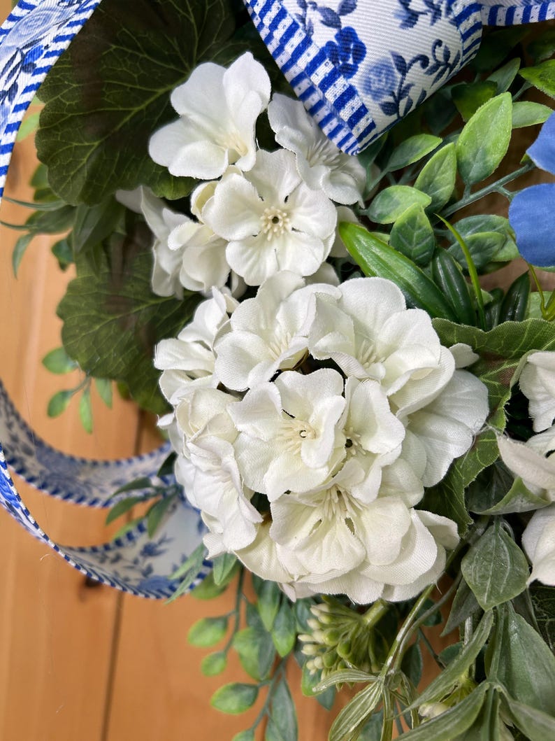 May include: A decorative wreath featuring clusters of white geranium flowers, green foliage, and a blue and white striped ribbon with a floral pattern. The arrangement includes various leaves and stems interspersed among the white blossoms.