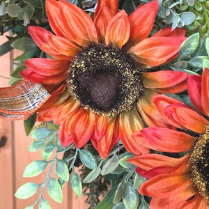 May include: A decorative wreath featuring large, vibrant orange and red sunflowers with dark brown centers dusted with yellow pollen. The sunflowers are surrounded by green foliage and accented with a plaid ribbon in shades of orange and brown.