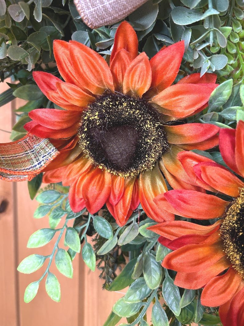 May include: A decorative wreath featuring large, vibrant orange and red sunflowers with dark brown centers dusted with yellow pollen. The sunflowers are surrounded by green foliage and accented with a plaid ribbon in shades of orange and brown.