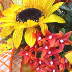 May include: A decorative floral arrangement featuring a large yellow sunflower with a dark brown center, clusters of small red star-shaped flowers with white centers, and green foliage. An orange and white plaid ribbon is woven through the flowers.