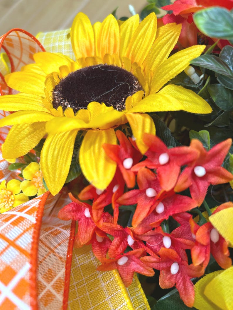 May include: A decorative floral arrangement featuring a large yellow sunflower with a dark brown center, clusters of small red star-shaped flowers with white centers, and green foliage. An orange and white plaid ribbon is woven through the flowers.