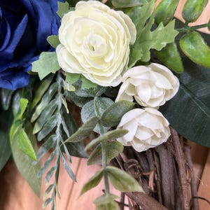Op de afbeelding: Een decoratieve krans met een grote donkerblauwe stoffen roos, twee kleinere cr&egrave;mekleurige stoffen roosjes en diverse groene kunstbladeren en stengels bevestigd op een basis van in elkaar gevlochten bruine natuurlijke takken en twijgen.