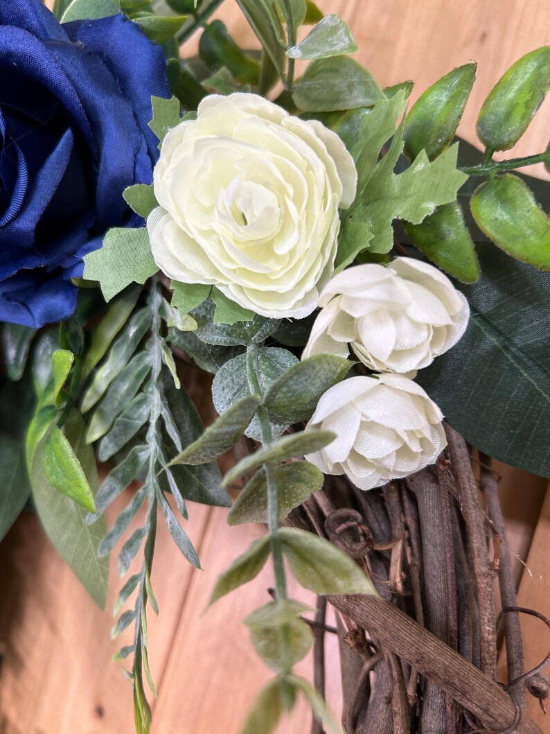 Op de afbeelding: Een decoratieve krans met een grote donkerblauwe stoffen roos, twee kleinere cr&egrave;mekleurige stoffen roosjes en diverse groene kunstbladeren en stengels bevestigd op een basis van in elkaar gevlochten bruine natuurlijke takken en twijgen.