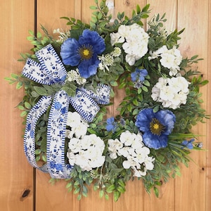 May include: A circular wreath featuring green foliage, white hydrangea clusters, and large blue poppy-like flowers. A decorative ribbon with a blue and white floral pattern and striped edges is tied to the left side, adding a contrasting accent.