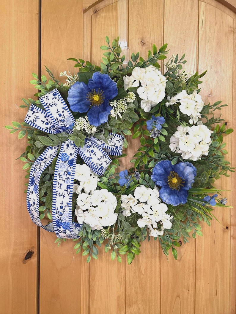 May include: A circular wreath featuring green foliage, white hydrangea clusters, and large blue poppy-like flowers. A decorative ribbon with a blue and white floral pattern and striped edges is tied to the left side, adding a contrasting accent.