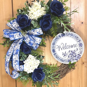 May include: A round grapevine wreath featuring deep navy blue roses, white hydrangea clusters, and green foliage. A large bow with a blue and white floral pattern is attached to the left side, and a circular sign with the word Welcome is on the right.