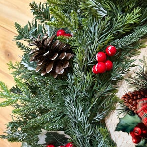 May include: A holiday wreath featuring artificial green cedar and pine branches with a light dusting of white frost. The arrangement includes clusters of bright red berries and brown pinecones, accented by green leaves and a plaid fabric element.