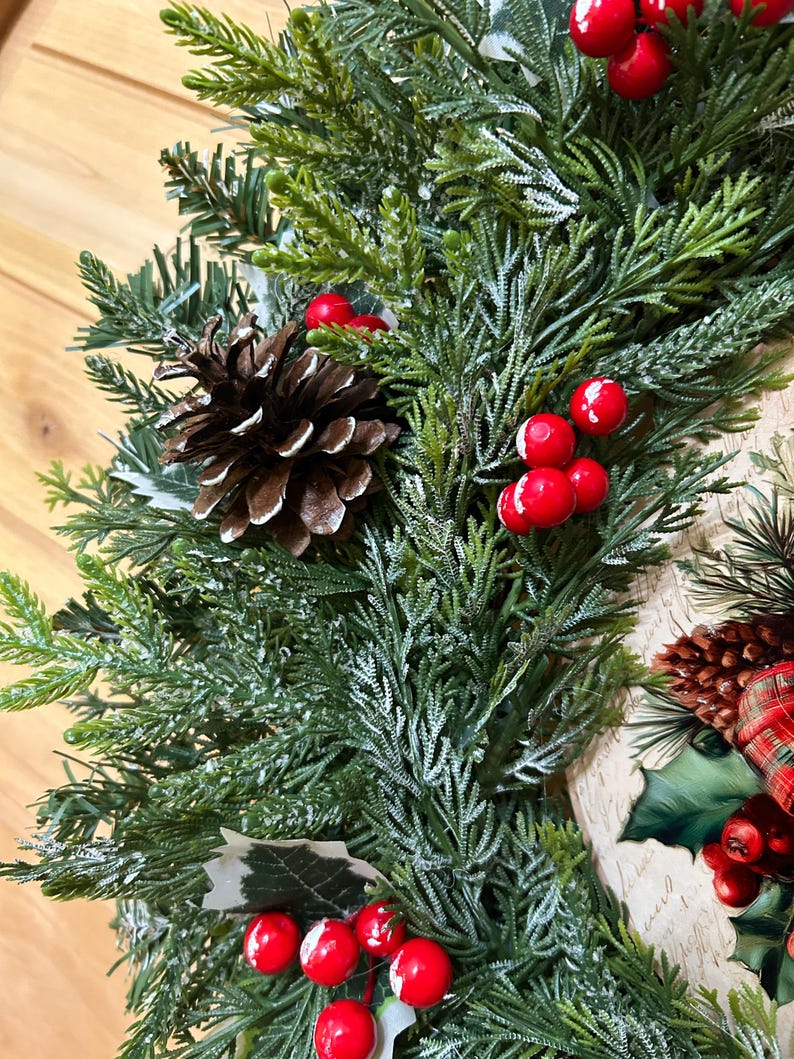 May include: A holiday wreath featuring artificial green cedar and pine branches with a light dusting of white frost. The arrangement includes clusters of bright red berries and brown pinecones, accented by green leaves and a plaid fabric element.