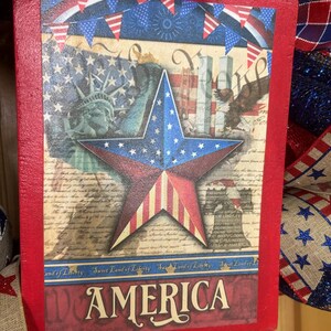 May include: A rectangular wooden sign with a red border featuring a patriotic collage of the Statue of Liberty, a bald eagle, the Liberty Bell, and a central star. The text reads Sweet Land of Liberty and AMERICA in a bold serif font.