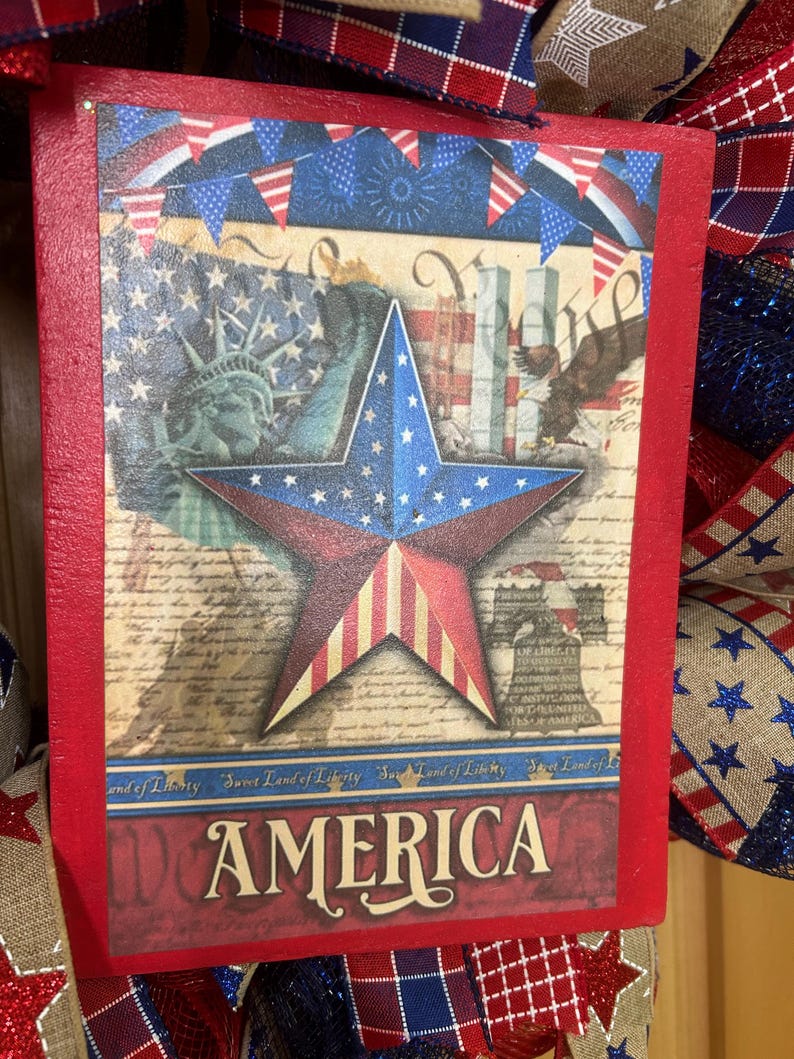 May include: A rectangular wooden sign with a red border featuring a patriotic collage of the Statue of Liberty, a bald eagle, the Liberty Bell, and a central star. The text reads Sweet Land of Liberty and AMERICA in a bold serif font.