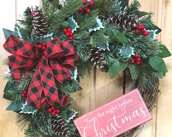 May include: A Christmas wreath featuring green pine needles, holly leaves, red berries, and brown pinecones. A large red and black buffalo plaid ribbon is attached to the left side. A rectangular red sign with white text reads 'Twas the night before Christmas'.