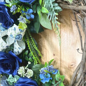 Op de afbeelding: Een decoratieve krans met diep marineblauwe rozen, kleinere lichtblauwe bloemen en groen blad op een basis van natuurlijke bruine takken. Een cr&egrave;mekleurig lint met een blauw bloemenpatroon is door het bloemstuk geweven.
