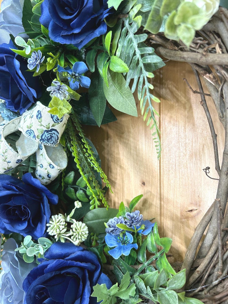 Op de afbeelding: Een decoratieve krans met diep marineblauwe rozen, kleinere lichtblauwe bloemen en groen blad op een basis van natuurlijke bruine takken. Een cr&egrave;mekleurig lint met een blauw bloemenpatroon is door het bloemstuk geweven.