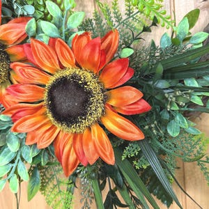 May include: A decorative wreath featuring large, vibrant orange sunflowers with dark brown centers and yellow pollen accents. The sunflowers are surrounded by a dense arrangement of various green foliage, including fern-like leaves, eucalyptus, and long, slender grass blades.