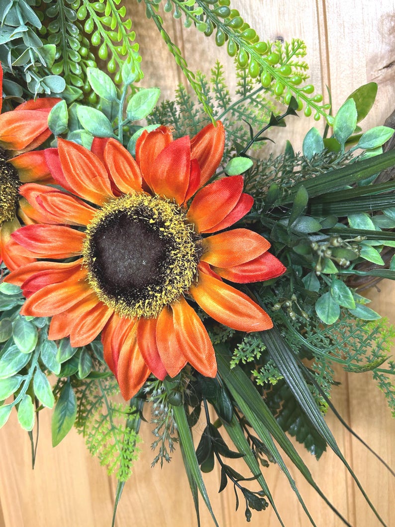 May include: A decorative wreath featuring large, vibrant orange sunflowers with dark brown centers and yellow pollen accents. The sunflowers are surrounded by a dense arrangement of various green foliage, including fern-like leaves, eucalyptus, and long, slender grass blades.