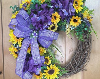 May include: A circular wreath featuring yellow sunflowers, deep purple hydrangea clusters, and green foliage arranged on a natural brown twig base. A large, multi-looped purple and white plaid ribbon is attached to the left side of the floral arrangement.