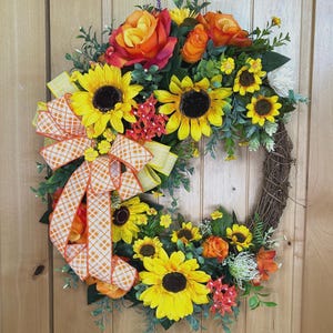 May include: A decorative wreath featuring bright yellow sunflowers, orange roses, and green foliage arranged on a brown twig base. A large, multi-layered bow with an orange and white checkered pattern is attached to the left side of the floral arrangement.
