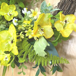 May include: A decorative wreath featuring bright yellow flowers with prominent white stamens and dark centers, interspersed with various green fern fronds and foliage. The floral arrangement is attached to a circular base made of woven brown twigs.