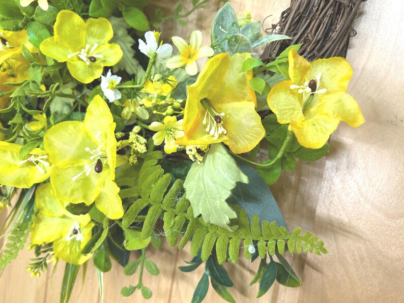 May include: A decorative wreath featuring bright yellow flowers with prominent white stamens and dark centers, interspersed with various green fern fronds and foliage. The floral arrangement is attached to a circular base made of woven brown twigs.