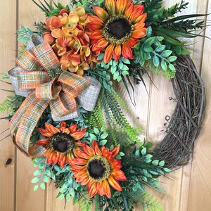 May include: A decorative wreath featuring three large orange and yellow sunflowers with dark brown centers, a cluster of orange hydrangea-like flowers, and various green foliage. A plaid ribbon in shades of orange, green, and tan is attached.