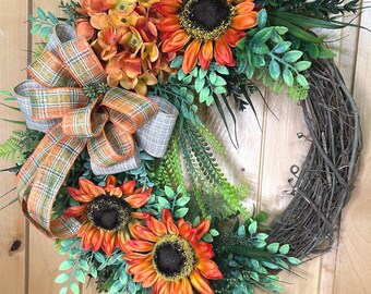 May include: A decorative wreath featuring three large orange and yellow sunflowers with dark brown centers, a cluster of orange hydrangea-like flowers, and various green foliage. A plaid ribbon in shades of orange, green, and tan is attached.