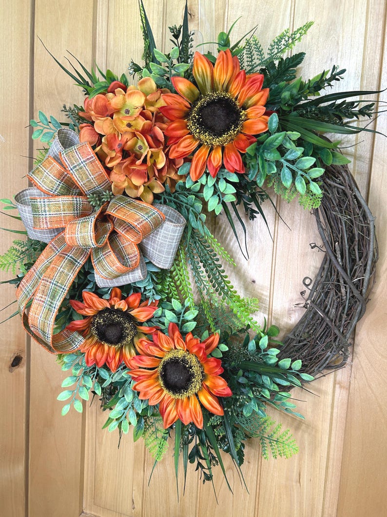 May include: A decorative wreath featuring three large orange and yellow sunflowers with dark brown centers, a cluster of orange hydrangea-like flowers, and various green foliage. A plaid ribbon in shades of orange, green, and tan is attached.