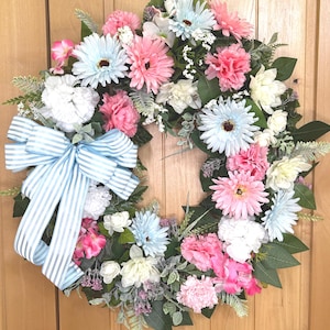 May include: A circular floral wreath featuring an assortment of artificial light blue, pink, and white flowers, including daisies and carnations, accented with green foliage and a large, light blue and white striped fabric bow on the left side.