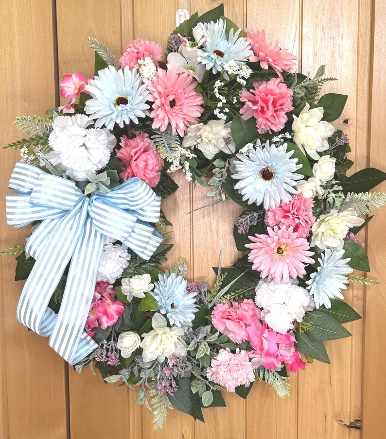 May include: A circular floral wreath featuring an assortment of artificial light blue, pink, and white flowers, including daisies and carnations, accented with green foliage and a large, light blue and white striped fabric bow on the left side.