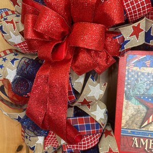 May include: A patriotic wreath featuring a large, glittery red bow as the centrepiece. The wreath is constructed from layers of blue mesh, hessian ribbon with white star patterns, red and blue plaid ribbon, and a rectangular sign depicting the Statue of Liberty.