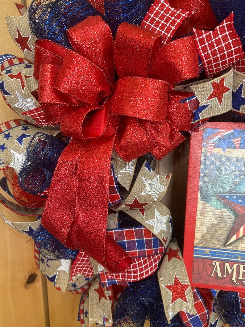 May include: A patriotic wreath featuring a large, glittery red bow as the centrepiece. The wreath is constructed from layers of blue mesh, hessian ribbon with white star patterns, red and blue plaid ribbon, and a rectangular sign depicting the Statue of Liberty.
