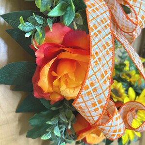May include: A decorative wreath featuring large orange and red roses, yellow sunflowers, and green foliage. A wide ribbon with an orange and white diamond grid pattern is tied in a bow, cascading down the center of the floral arrangement.