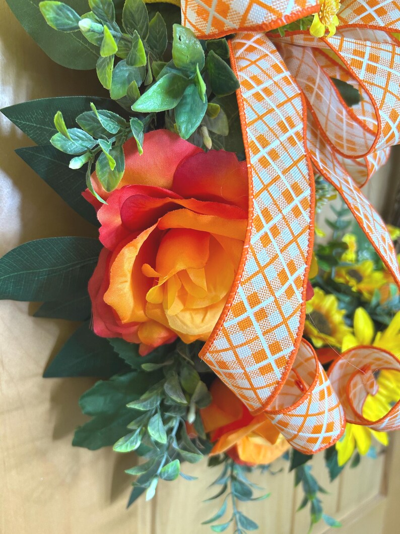 May include: A decorative wreath featuring large orange and red roses, yellow sunflowers, and green foliage. A wide ribbon with an orange and white diamond grid pattern is tied in a bow, cascading down the center of the floral arrangement.