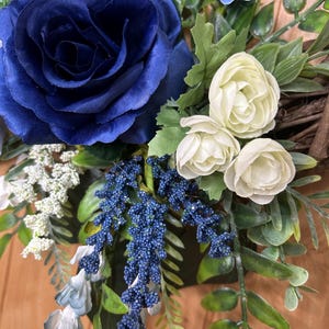 Op de afbeelding: Een decoratieve krans met een grote donkerblauwe roos, kleinere cr&egrave;mekleurige roosjes, trossen kleine blauwe besjes, witte vulbloemen en groen blad, gerangschikt op een basis van bruine takjes met diverse texturen.
