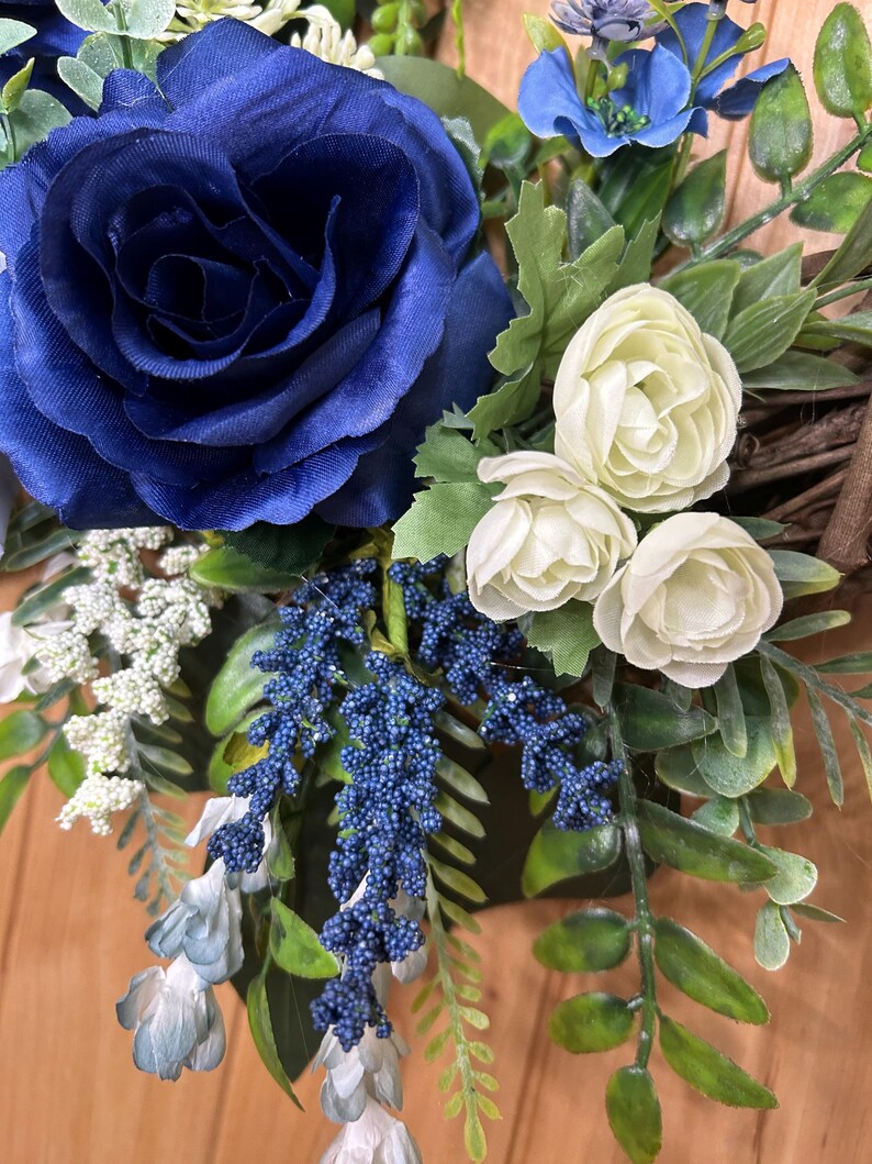 Op de afbeelding: Een decoratieve krans met een grote donkerblauwe roos, kleinere cr&egrave;mekleurige roosjes, trossen kleine blauwe besjes, witte vulbloemen en groen blad, gerangschikt op een basis van bruine takjes met diverse texturen.