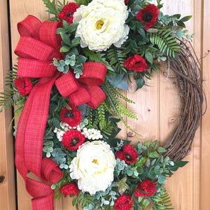 May include: A decorative wreath featuring a natural brown grapevine base adorned with large white peony-like flowers, vibrant red poppies, green foliage, and small white berries, finished with a prominent red textured fabric bow on the left side.