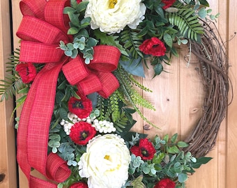 May include: A decorative wreath featuring a natural brown grapevine base adorned with large white peony-like flowers, vibrant red poppies, green foliage, and small white berries, finished with a prominent red textured fabric bow on the left side.