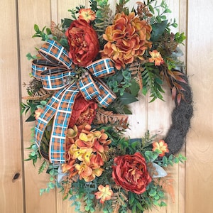 May include: A circular wreath featuring artificial orange and red flowers, green foliage, and brown fern accents. A large decorative bow with a blue, orange, and white plaid pattern is attached to the left side of the wreath, which is built on a dark brown base.