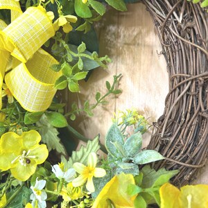 May include: A decorative wreath featuring a natural brown twig base adorned with bright yellow flowers, green foliage, and a yellow and white checkered ribbon. The arrangement includes various small blossoms and leaves attached to the circular frame.