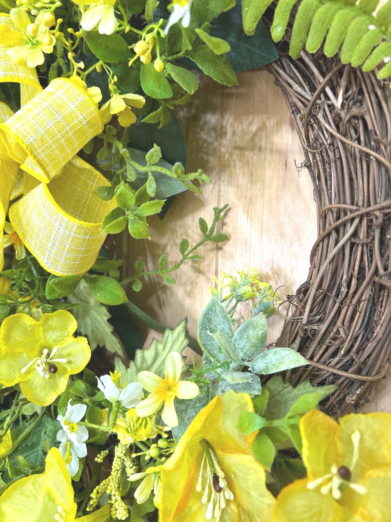 May include: A decorative wreath featuring a natural brown twig base adorned with bright yellow flowers, green foliage, and a yellow and white checkered ribbon. The arrangement includes various small blossoms and leaves attached to the circular frame.