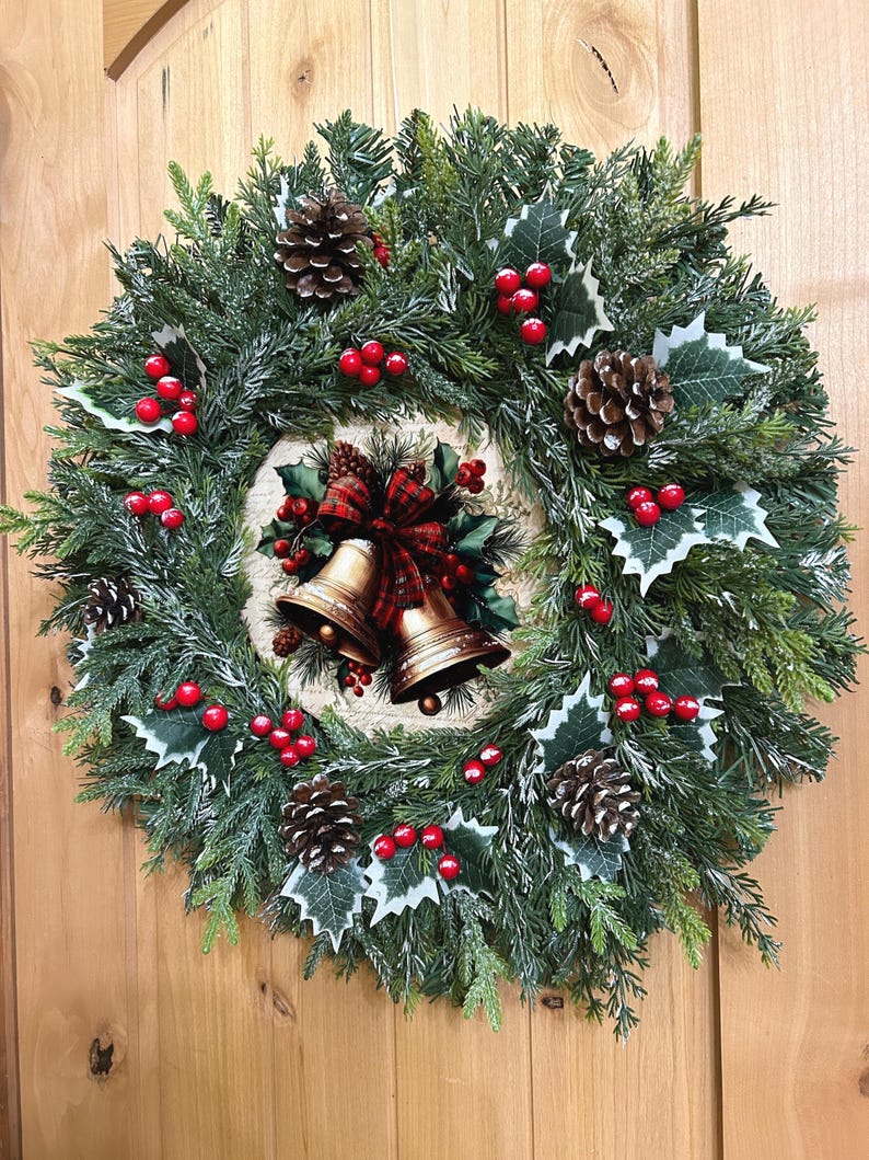 May include: A circular holiday wreath featuring green pine needles, brown pinecones, and clusters of bright red berries. The center displays a decorative circular plaque showing two gold-colored bells tied with a red and black plaid ribbon.