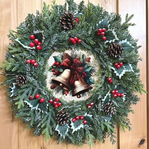 May include: A circular wreath made of green pine needles and frosted branches, decorated with brown pinecones, red berries, and green holly leaves. The center features a round plaque displaying two gold bells tied with a red and green plaid ribbon.