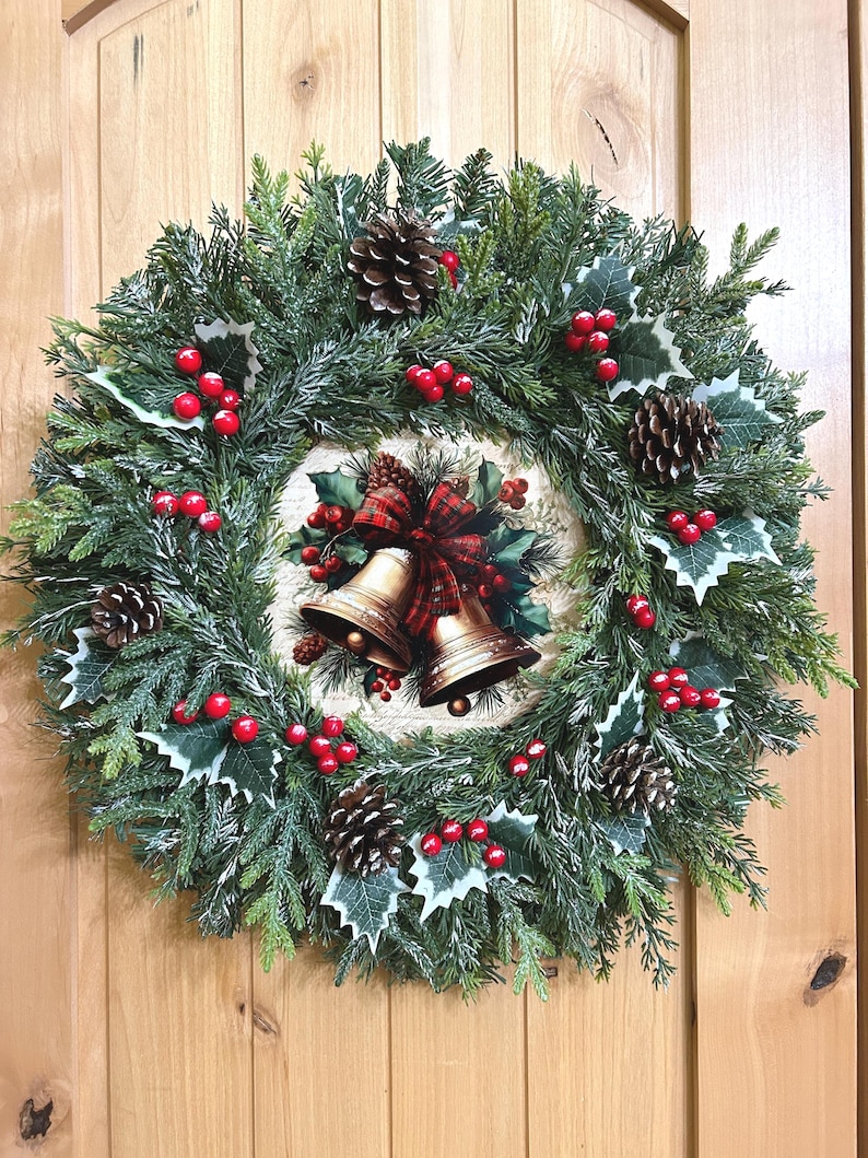 May include: A circular wreath made of green pine needles and frosted branches, decorated with brown pinecones, red berries, and green holly leaves. The center features a round plaque displaying two gold bells tied with a red and green plaid ribbon.