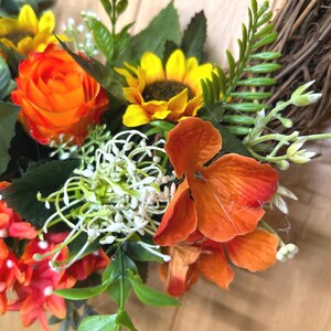 May include: A decorative wreath featuring a woven brown twig base adorned with artificial sunflowers, vibrant orange roses, small red star-shaped flowers, white filler blossoms, and green foliage. The arrangement displays a mix of warm colors.