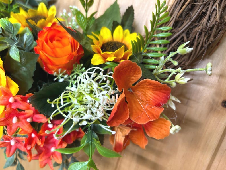 May include: A decorative wreath featuring a woven brown twig base adorned with artificial sunflowers, vibrant orange roses, small red star-shaped flowers, white filler blossoms, and green foliage. The arrangement displays a mix of warm colors.