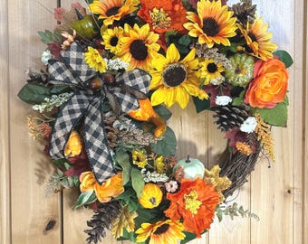 May include: A decorative wreath featuring yellow sunflowers, orange flowers, small green and orange pumpkins, brown pinecones, and autumn-coloured leaves. A black and white buffalo plaid ribbon bow is attached to the left side of the circular arrangement.